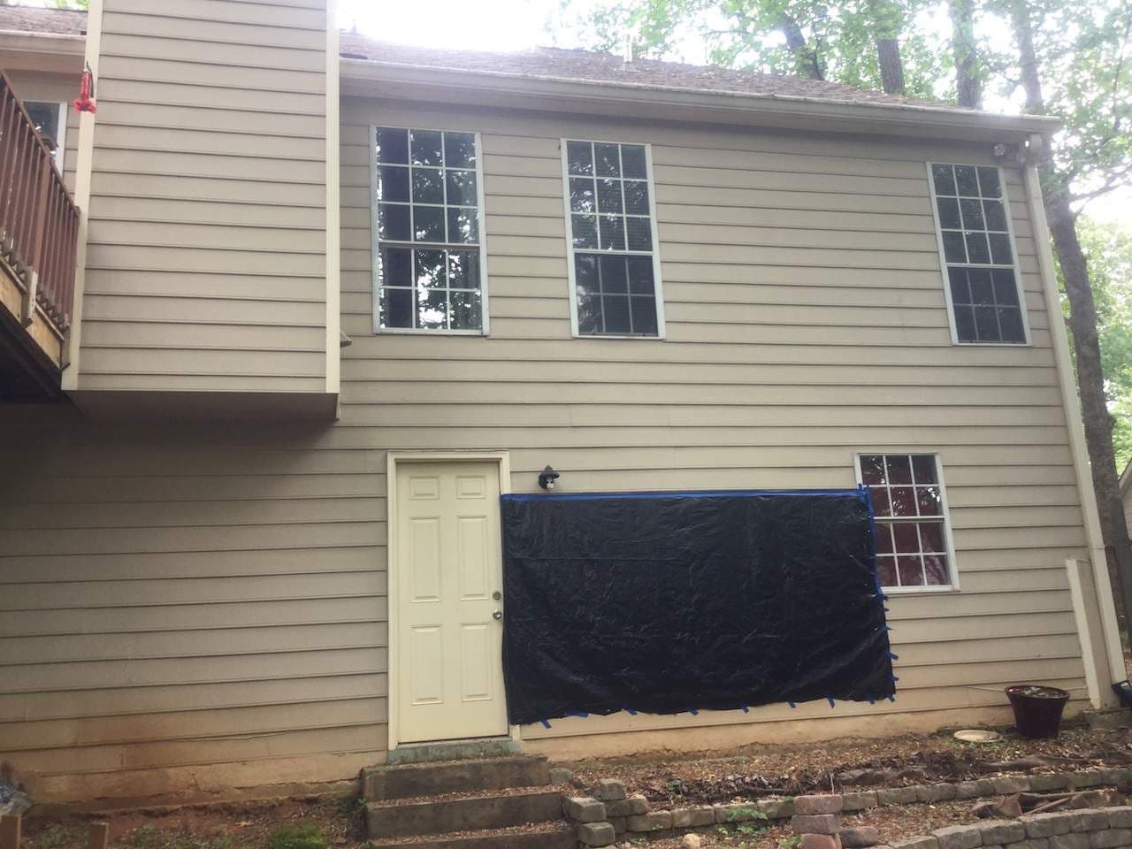 Two-story house exterior with beige siding, three large windows, a white door, and a section of siding covered by black plastic sheeting, suggesting ongoing repairs.