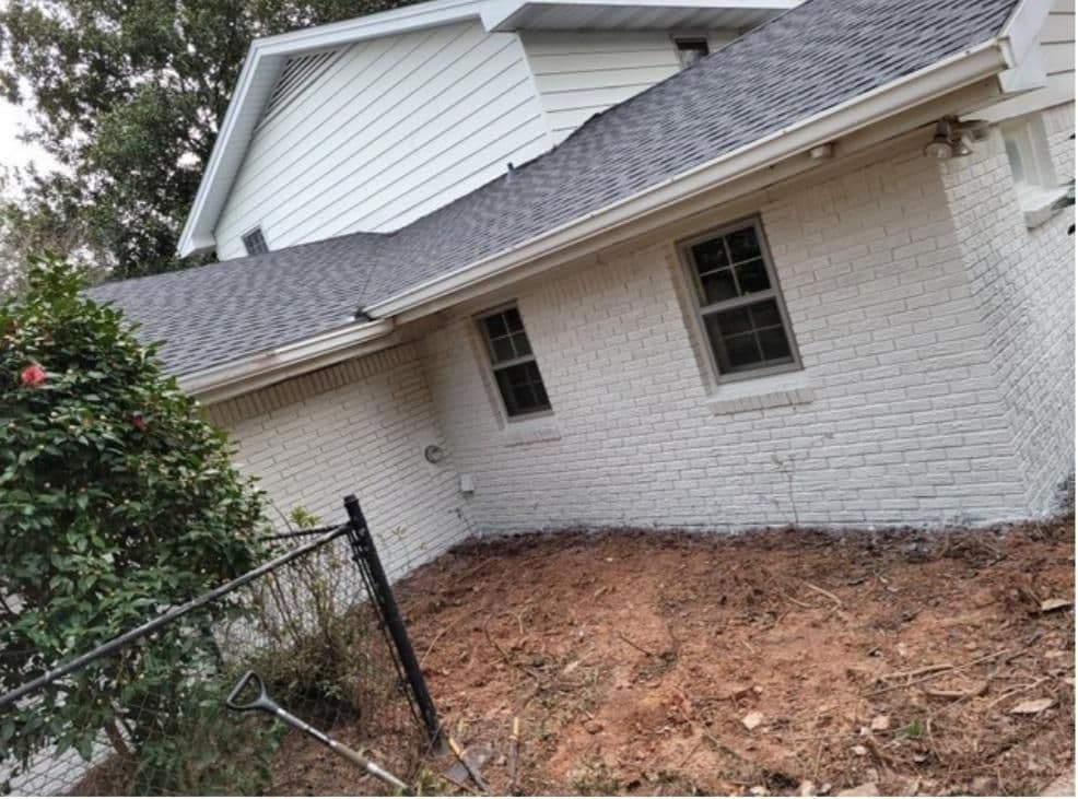 White brick house with two windows, gray shingle roof, and an adjacent bare dirt area. A shovel and a bush lean against a chain-link fence.