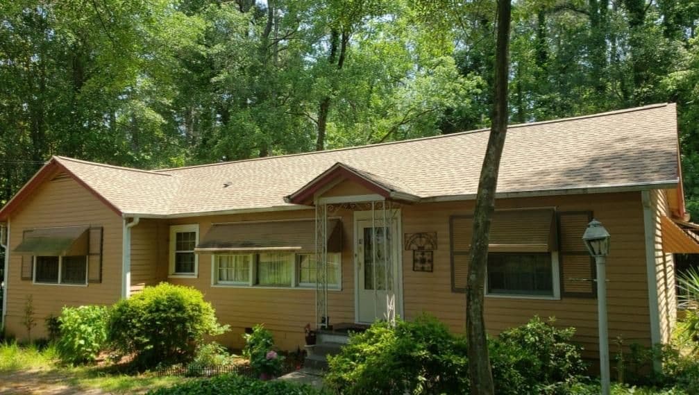 Cozy, light-brown house with a gabled roof, surrounded by lush greenery and trees. There's a pathway leading to the front door, evoking a welcoming feel.