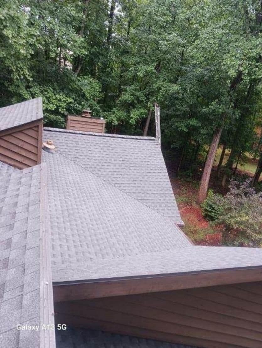 View from above of a gray shingled rooftop amidst a lush forest backdrop. The trees are dense and green, creating a serene and natural setting.