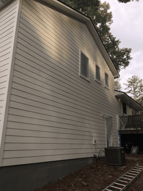 Side view of a beige house with horizontal siding, three covered windows, and an outdoor air conditioning unit. A metal ladder is on the ground, and the scene is calm.