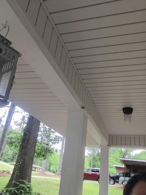 Covered porch with white ceiling and columns, a hanging lantern, and a light fixture. Background shows trees, grass, and parked cars, evoking a tranquil suburban feel.