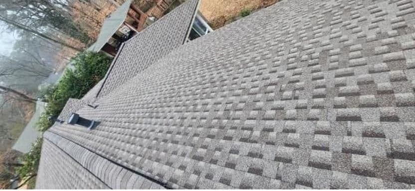 View of a gray shingle roof with a textured pattern on a house surrounded by trees. A chimney and foliage are visible in the background.
