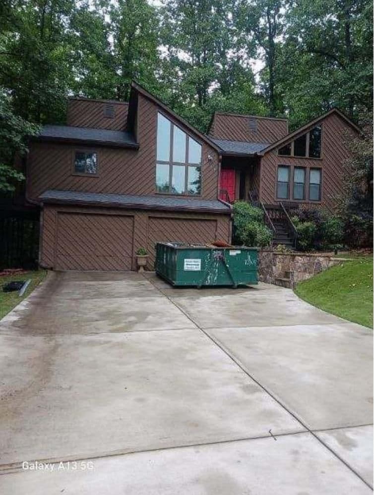 Modern brown house with large windows and red door, surrounded by trees. A green dumpster sits on the wet driveway, suggesting renovation work.
