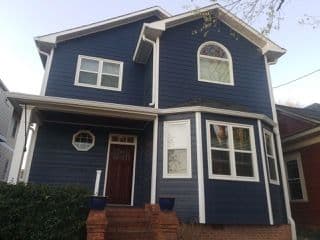 A two-story house with navy blue siding, white trim, and various windows, including a bay window. Brick steps lead to a red front door.