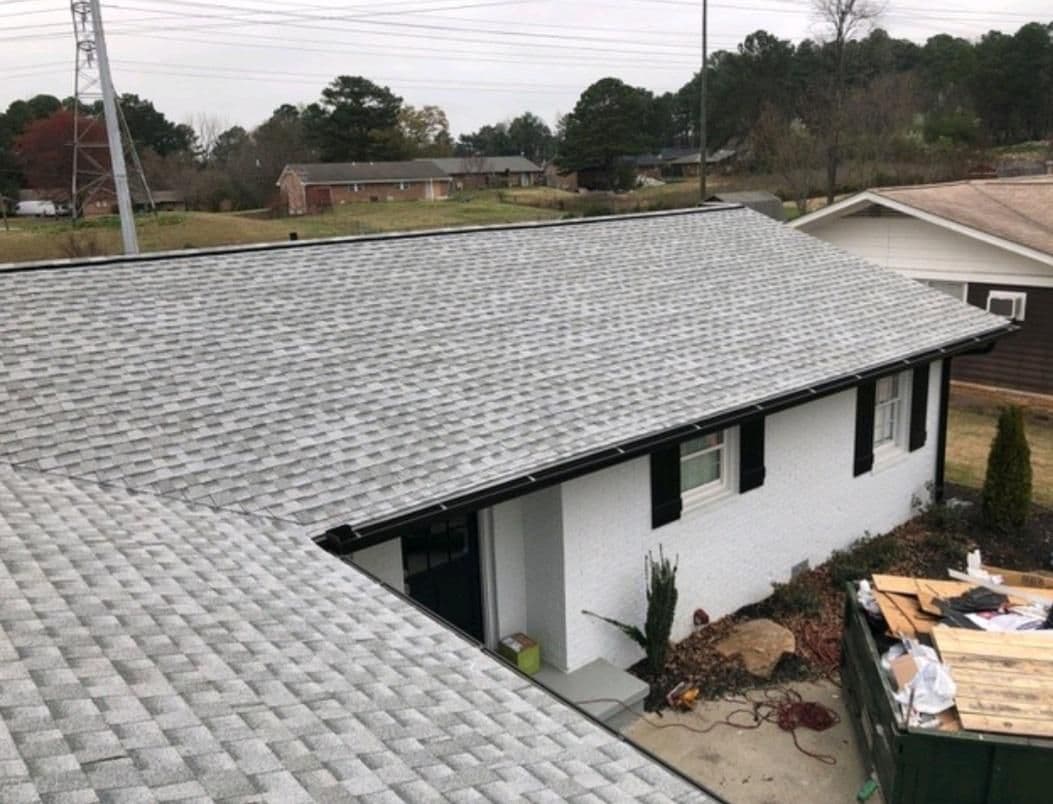 A newly shingled gray roof tops a white brick house with black shutters. In the yard, there's a green dumpster filled with construction debris.