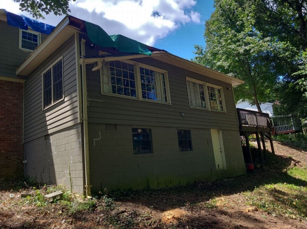 A house with damaged siding and a tarp on the roof, indicating repair. Surrounded by trees, the setting feels neglected yet peaceful.
