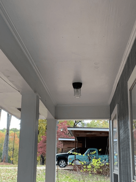 View of a covered porch with a hanging light fixture. A pickup truck is visible outside next to autumn trees, creating a calm, rural atmosphere.