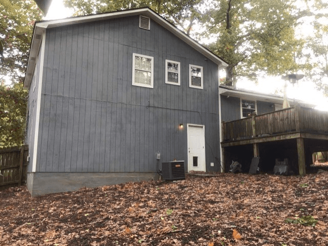 A gray, two-story house with three small windows and a white door on a sloped yard covered in fallen leaves. A wooden deck extends from the side.
