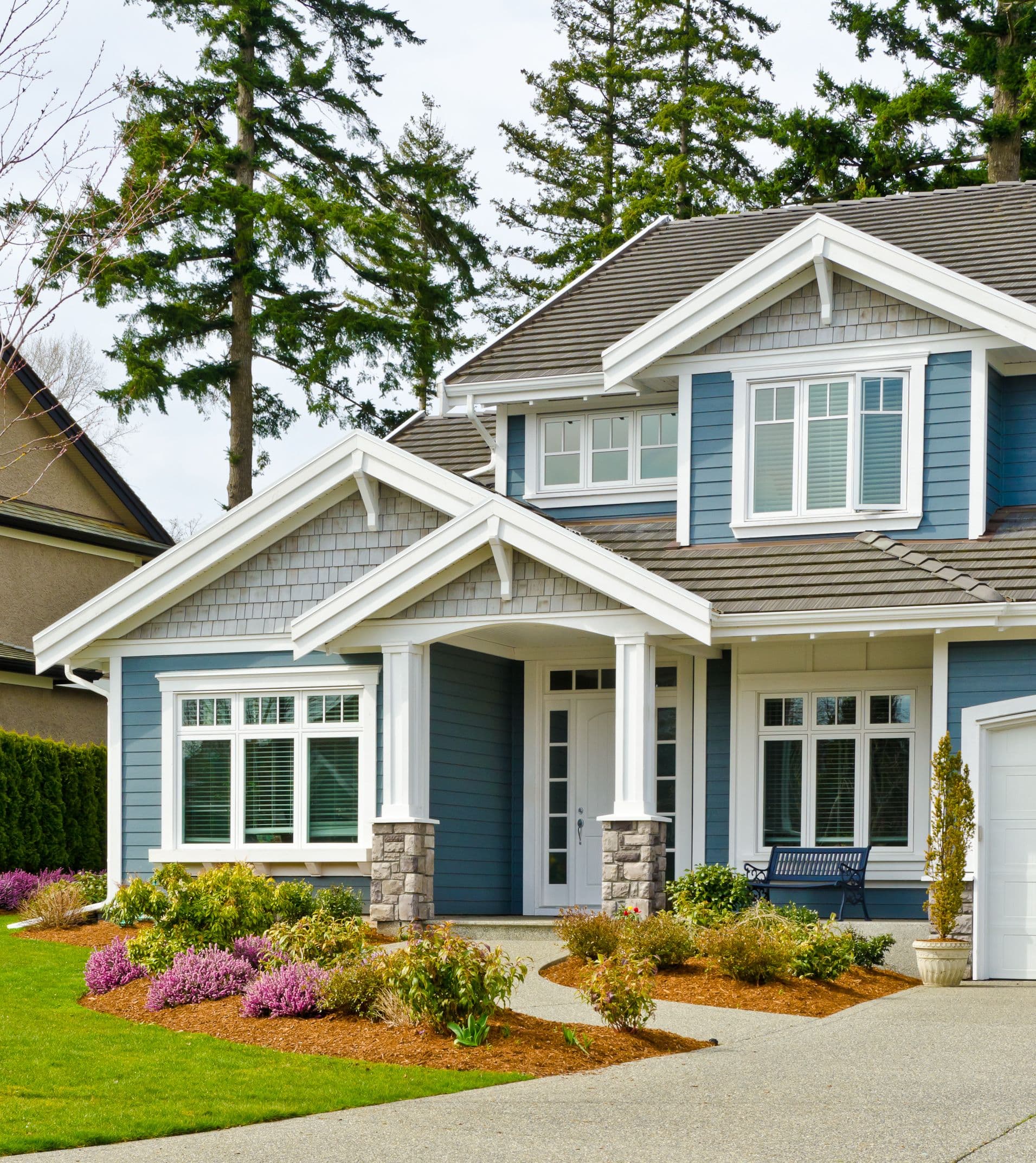 A modern two-story house with blue siding and white trim, surrounded by lush green lawn and colorful shrubs. The scene is peaceful and inviting.