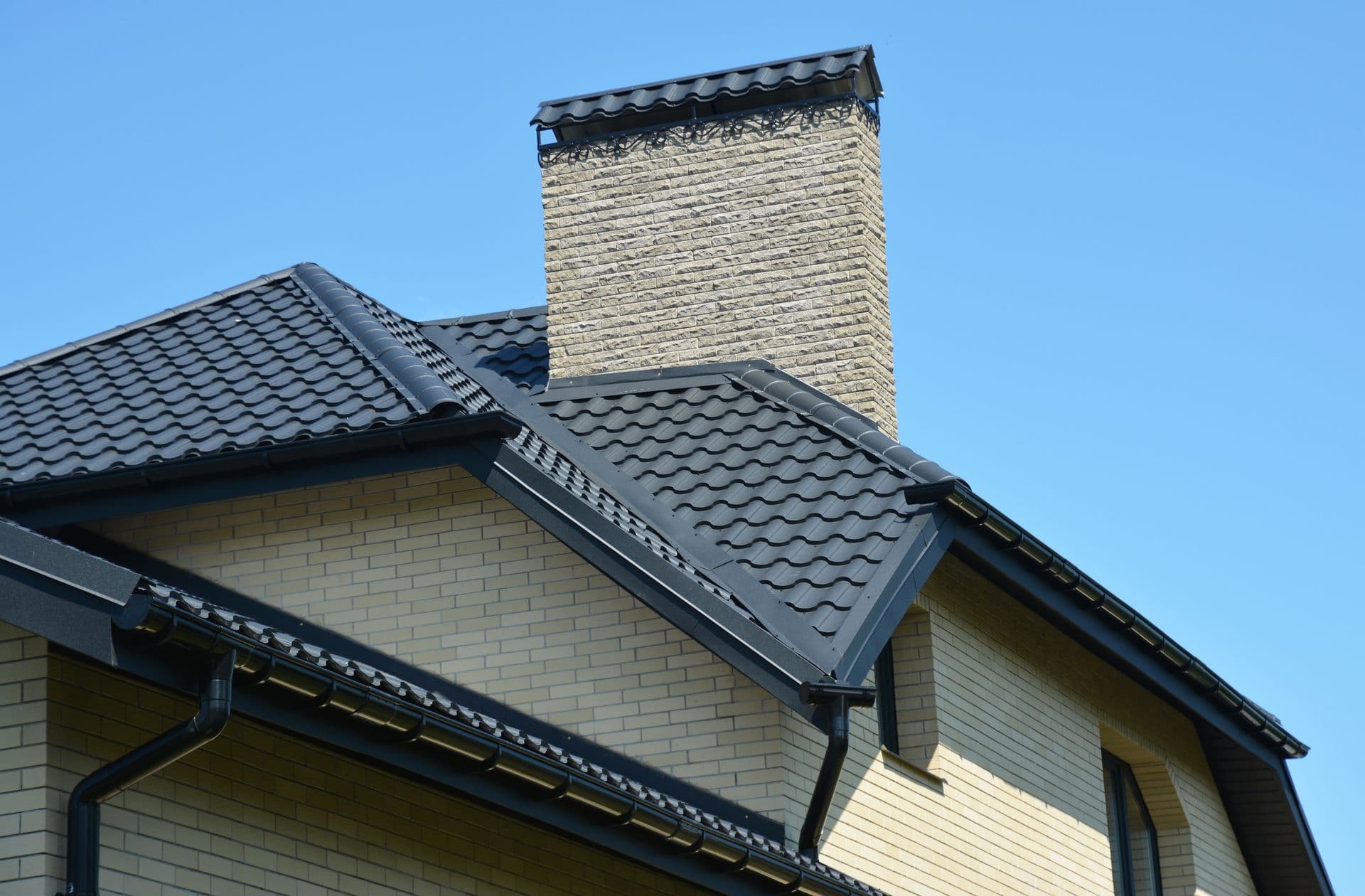 Brick house with a gabled roof and black shingles under a clear blue sky. Prominent chimney and sleek design convey modern elegance.