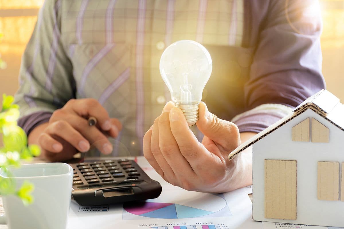 A person holds a glowing light bulb, symbolizing ideas, sits with a calculator and paper house model. Sunlight illuminates the workspace, suggesting innovation.