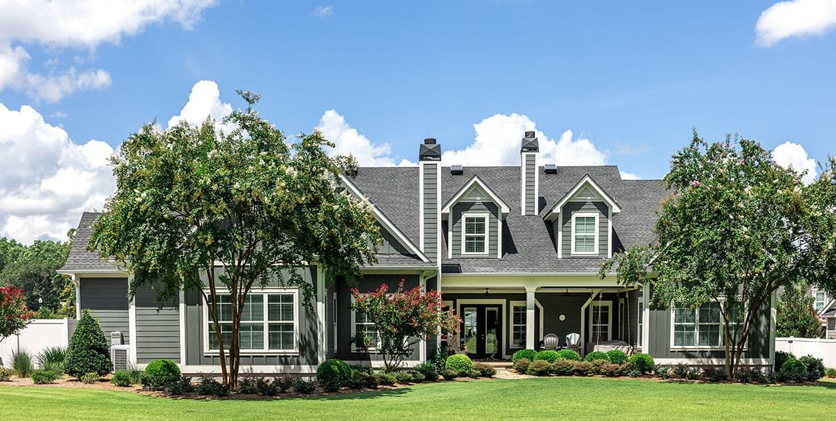 A charming gray house with white trim, dormer windows, and a porch is surrounded by lush green grass and trees under a bright blue sky with clouds.