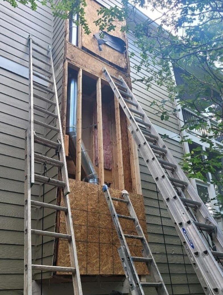 Two ladders lean against a house, reaching a wooden structure under construction. Exposed pipes are visible, with tree branches overhead.