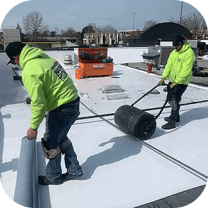 Two workers in bright green hoodies, on a flat white rooftop, are laying insulation material. The scene conveys teamwork and industrial work.