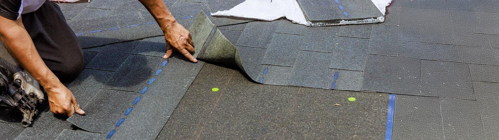 A person installing dark asphalt roof shingles, aligning them carefully. The image conveys precision and careful craftsmanship.