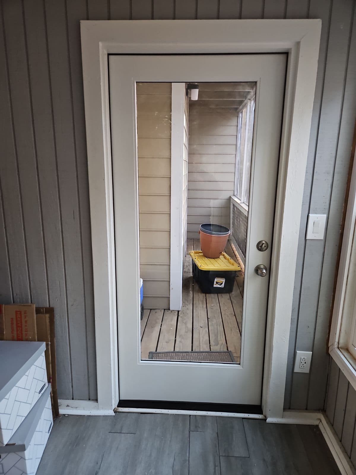 A glass door with a wooden frame opens to a small porch with wooden planks. A large pot sits on a yellow container, creating a simple, cozy setting.