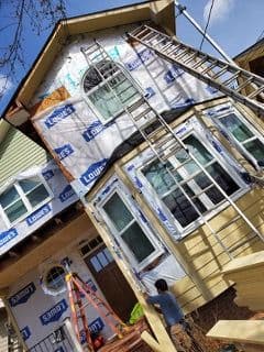 A person works on the exterior renovation of a house, using ladders and wearing blue overalls. The facade is partially covered with Lowe's branded wrap.
