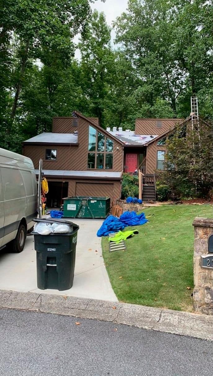 A modern, multi-level brown house with large windows is surrounded by lush trees. A ladder leans against the house, and blue tarps cover the ground. A white van is parked in the driveway beside a trash can.