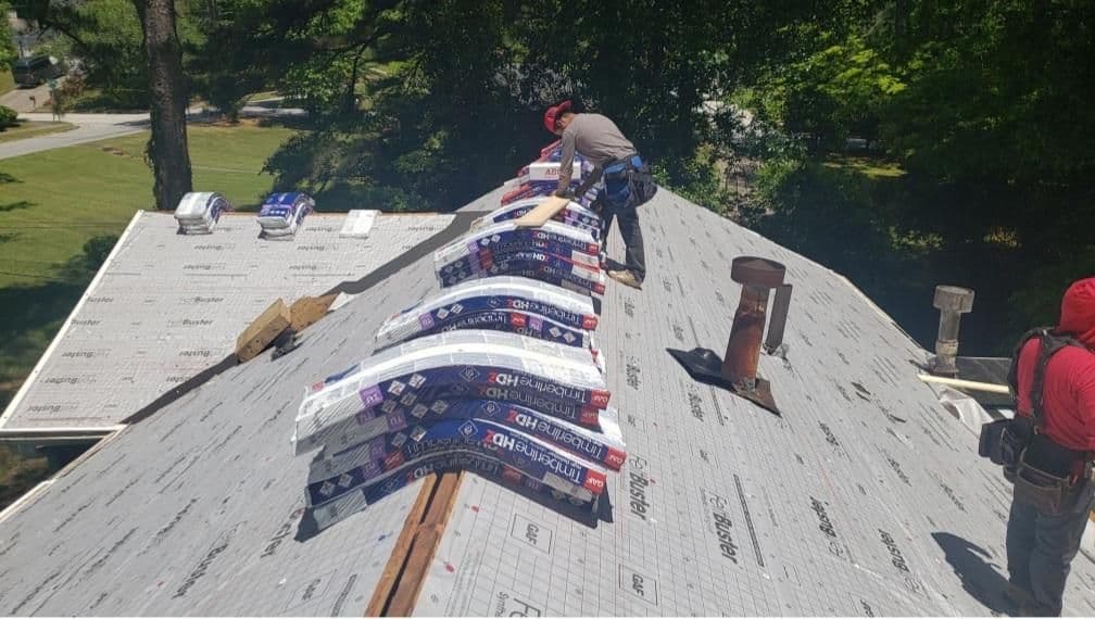 A roofer in a red helmet arranges stacks of shingles on a gray roof under bright sunlight. Another worker stands nearby, surrounded by lush trees.