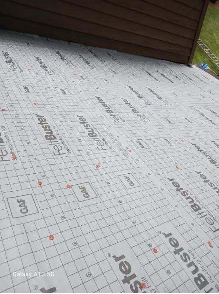 Roof underlayment covered with GAF FeltBuster sheets, displaying grid patterns and logos. The scene conveys a sense of construction or renovation.