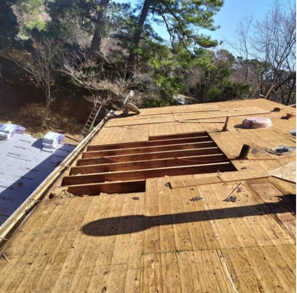 Partially constructed wooden roof with an exposed section revealing beams, surrounded by trees. A ladder leans against the structure.