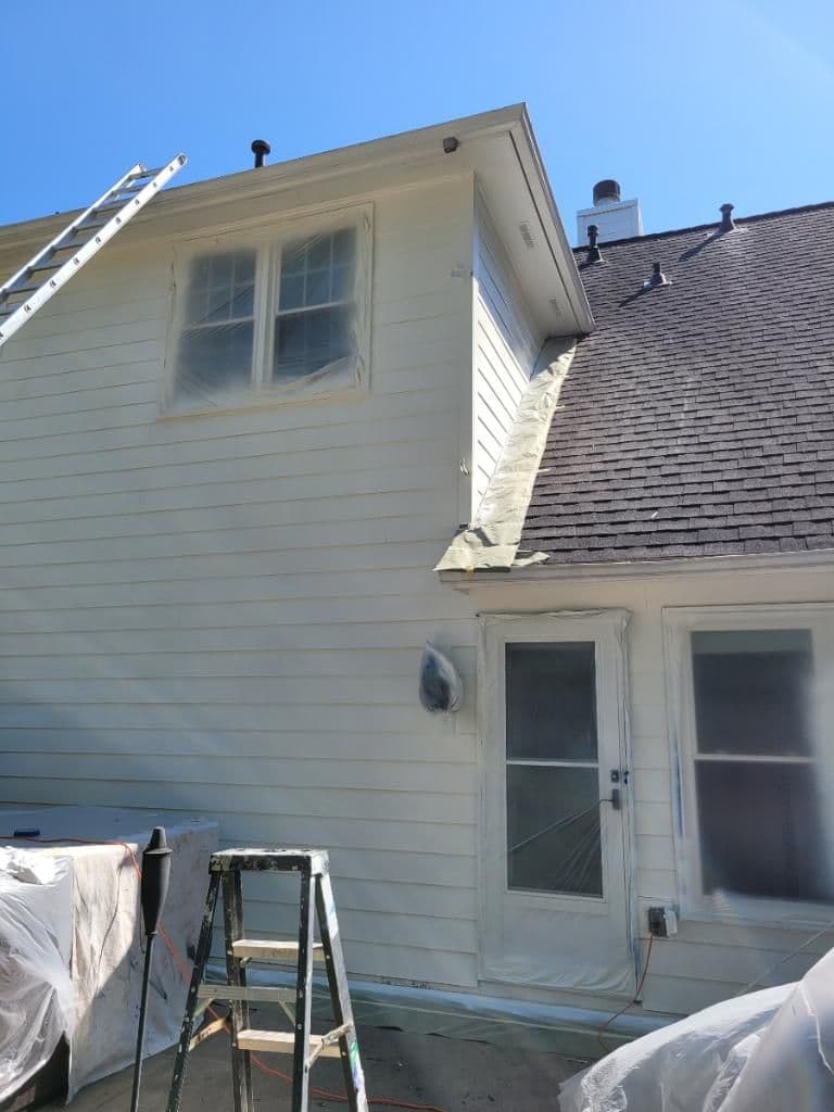 House exterior prepared for painting, with windows and door covered in protective plastic. A ladder leans against the white siding under a clear blue sky.