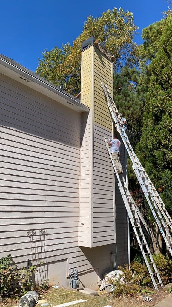 Two workers on ladders paint a beige chimney attached to a house, surrounded by trees under a clear blue sky. The scene is calm and focused.