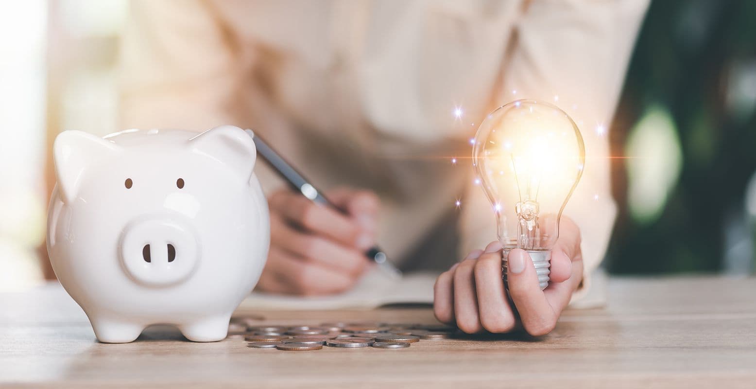 A person holds a glowing light bulb beside a white piggy bank on a table with coins. They write in a notebook, symbolizing financial ideas and planning.