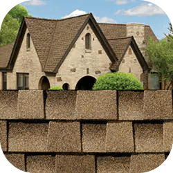 A stone house with a steep brown shingle roof is partially obscured by a stack of shingles. Trees and a blue sky with clouds are in the background.