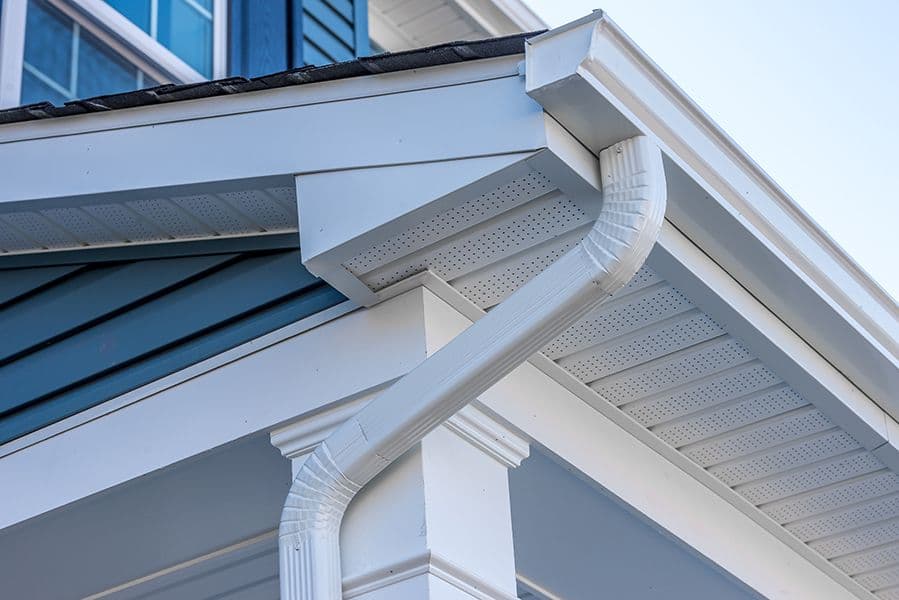 Close-up of a house roof corner showing a white downspout and gutter. The siding is blue with white trim, emphasizing a clean, modern design.