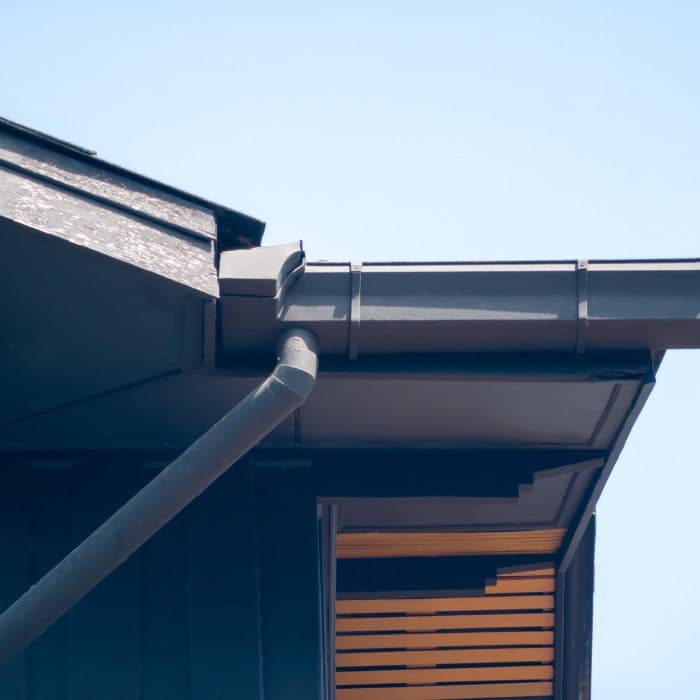 Close-up of a modern roof with a drainage system, showing grey gutters and downspouts against a clear blue sky. The structure appears sleek and well-maintained.