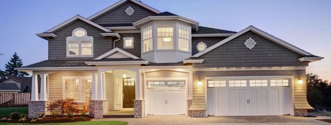 A modern two-story house with illuminated windows, a welcoming front porch, and a three-car garage. The exterior features beige siding and stone accents, against a twilight sky.