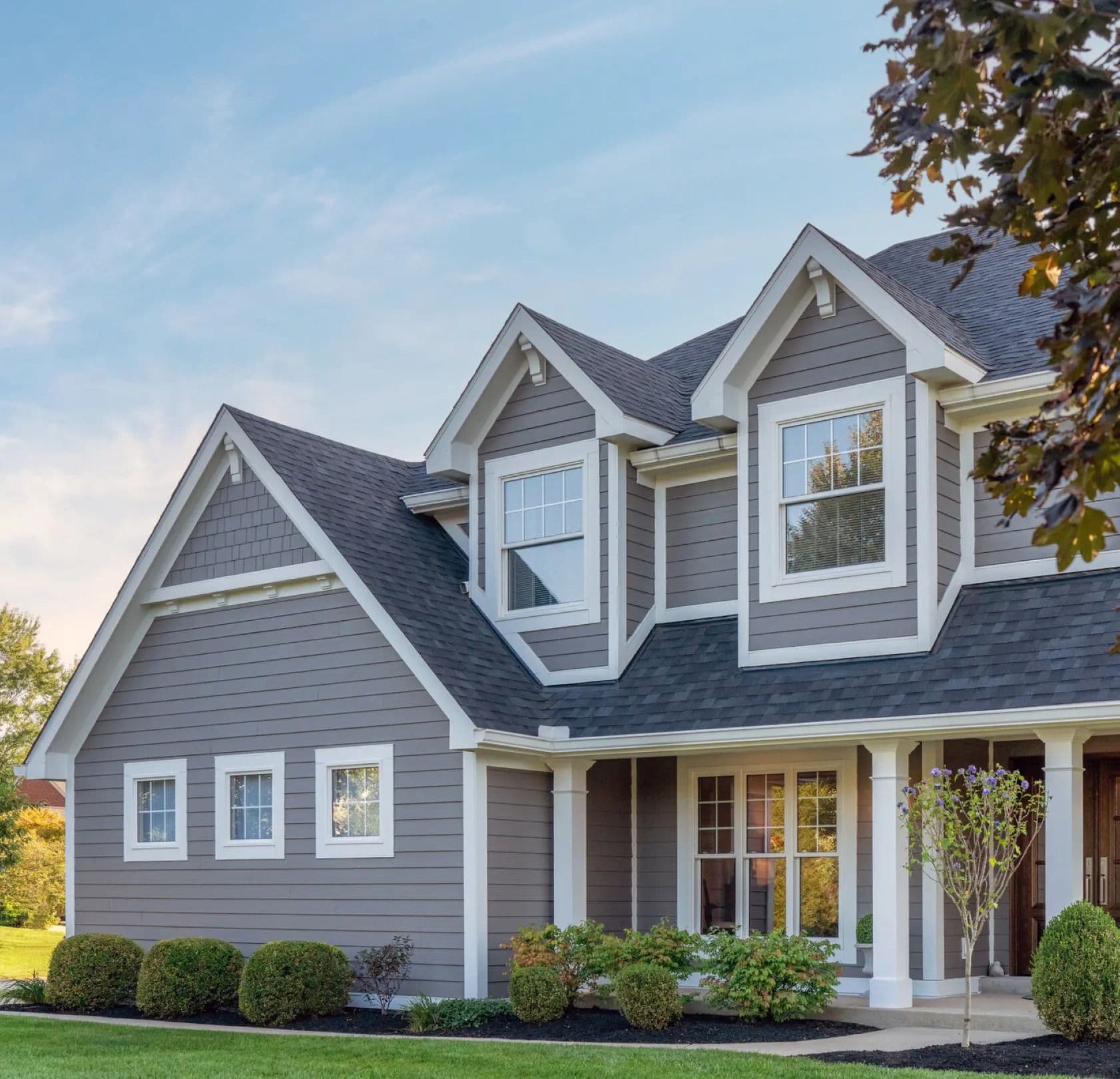 Two-story house with gray siding, white trim, and gabled roof. Neat shrubs and green lawn in front, under a clear blue sky with light clouds.