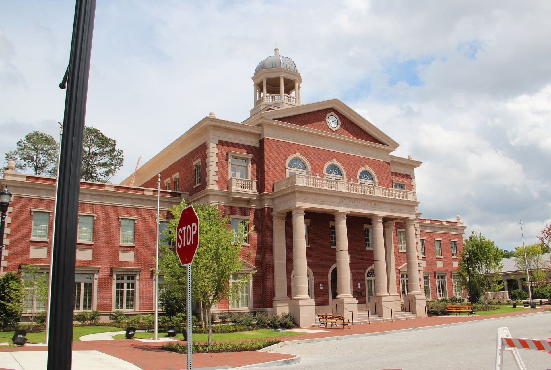 Historic red brick building with tall columns and a clock tower. A stop sign is in front, surrounded by greenery under a partly cloudy sky.