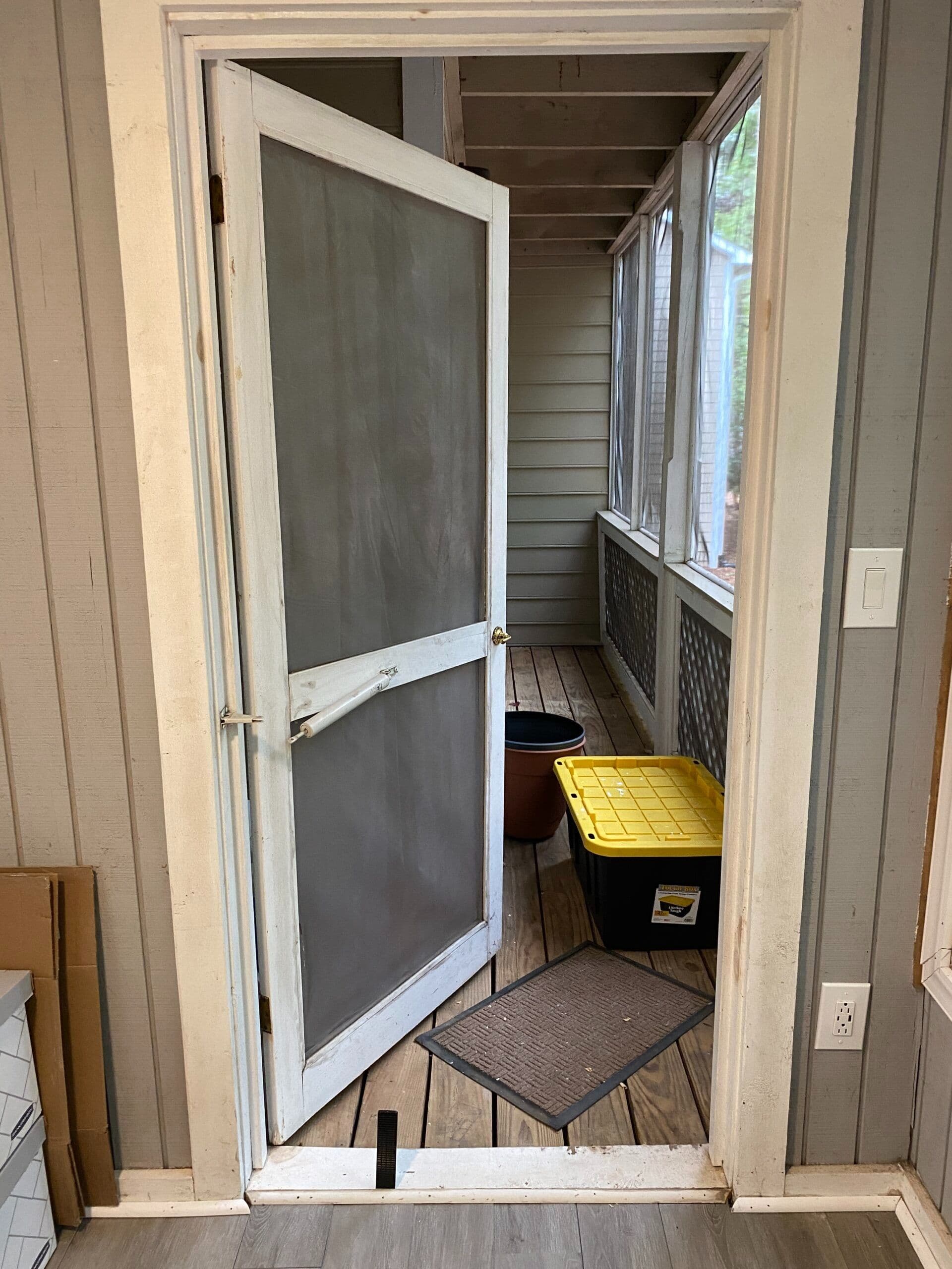 A white screen door slightly ajar leads to a narrow, enclosed porch with wooden flooring. A yellow-lidded plastic bin and a mat are visible, conveying a casual, tidy space.