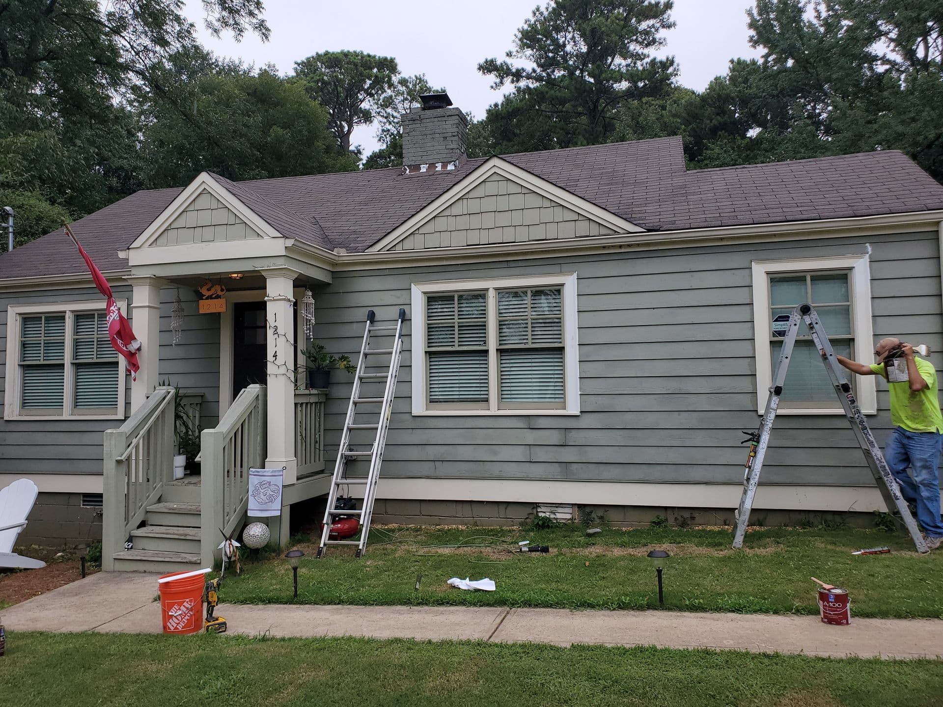 A gray wooden house under renovation, featuring ladders and paint cans. A worker in a yellow shirt paints near a window. Green lawn and trees in the background.