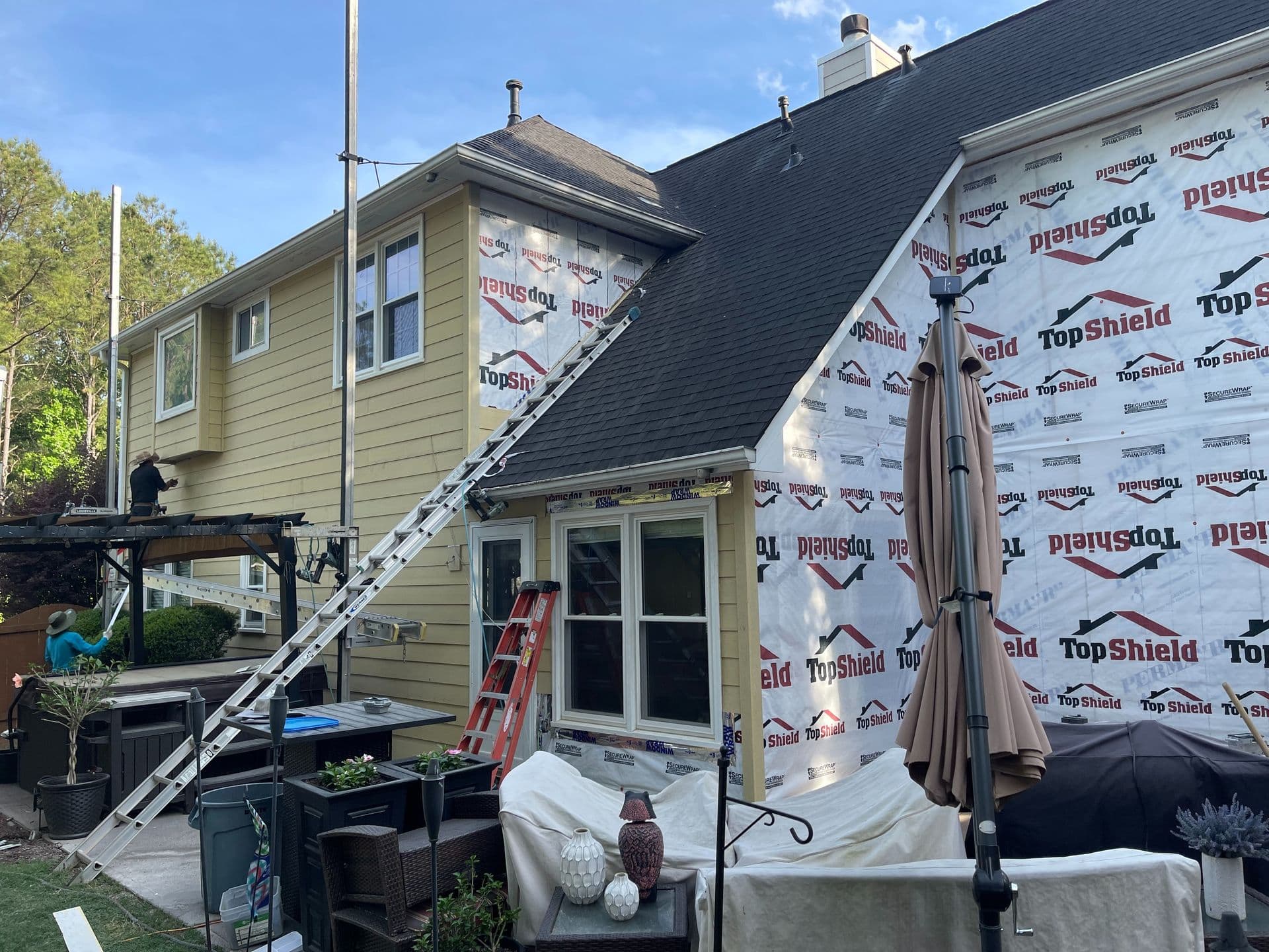 House under renovation with TopShield wrap, ladders, and scaffolding visible. Yellow siding halfway completed. Patio furniture covered below, clear sky above.
