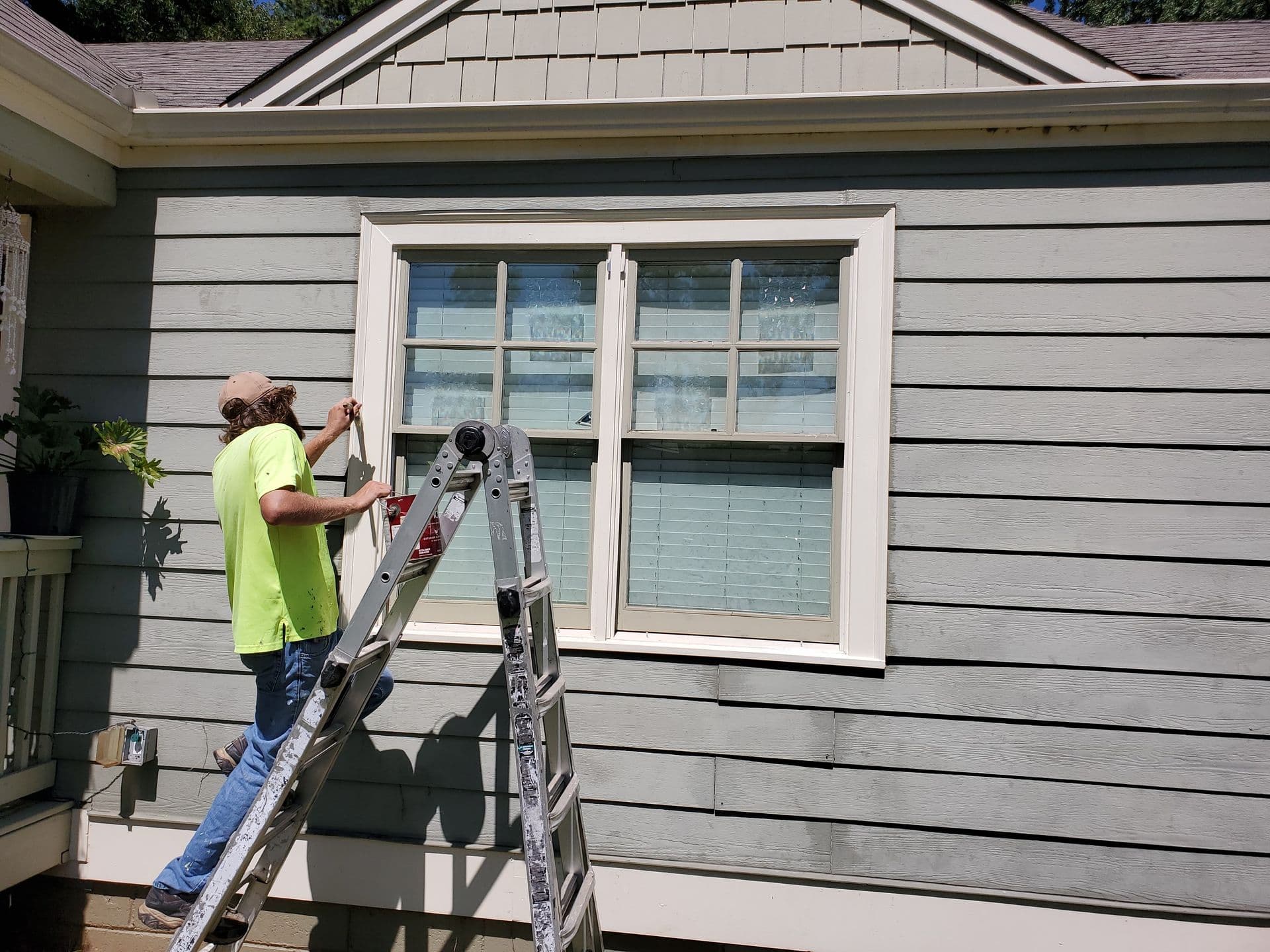 A person on a ladder paints the window trim of a light grey house. Bright sunlight accentuates the modern siding, creating a focused, industrious mood.