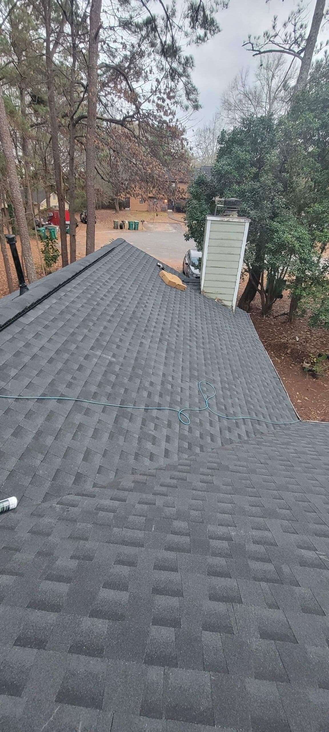 A brick house with a black roof is surrounded by tall trees under a cloudy sky. A light by the door illuminates a dumpster filled with construction debris.