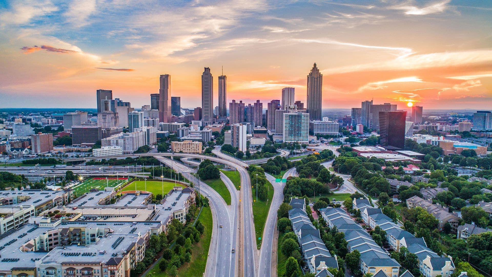 Aerial view of a city skyline at sunset with tall buildings and a prominent highway leading into the city. Vibrant sky colors and lush greenery create a serene atmosphere.