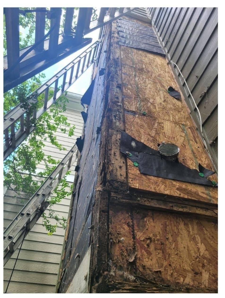Close-up view of a building corner under renovation, showing exposed wood and black tar paper. Two ladders lean against the structure amid trees.