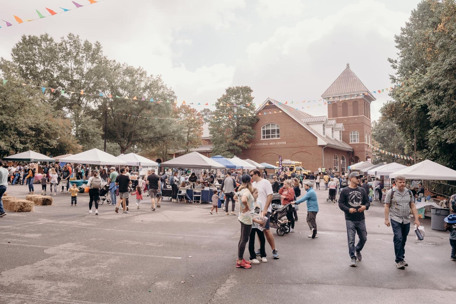 Outdoor community festival with people mingling near tents. Bunting hangs overhead, the atmosphere is lively and social. Red brick building backdrop.