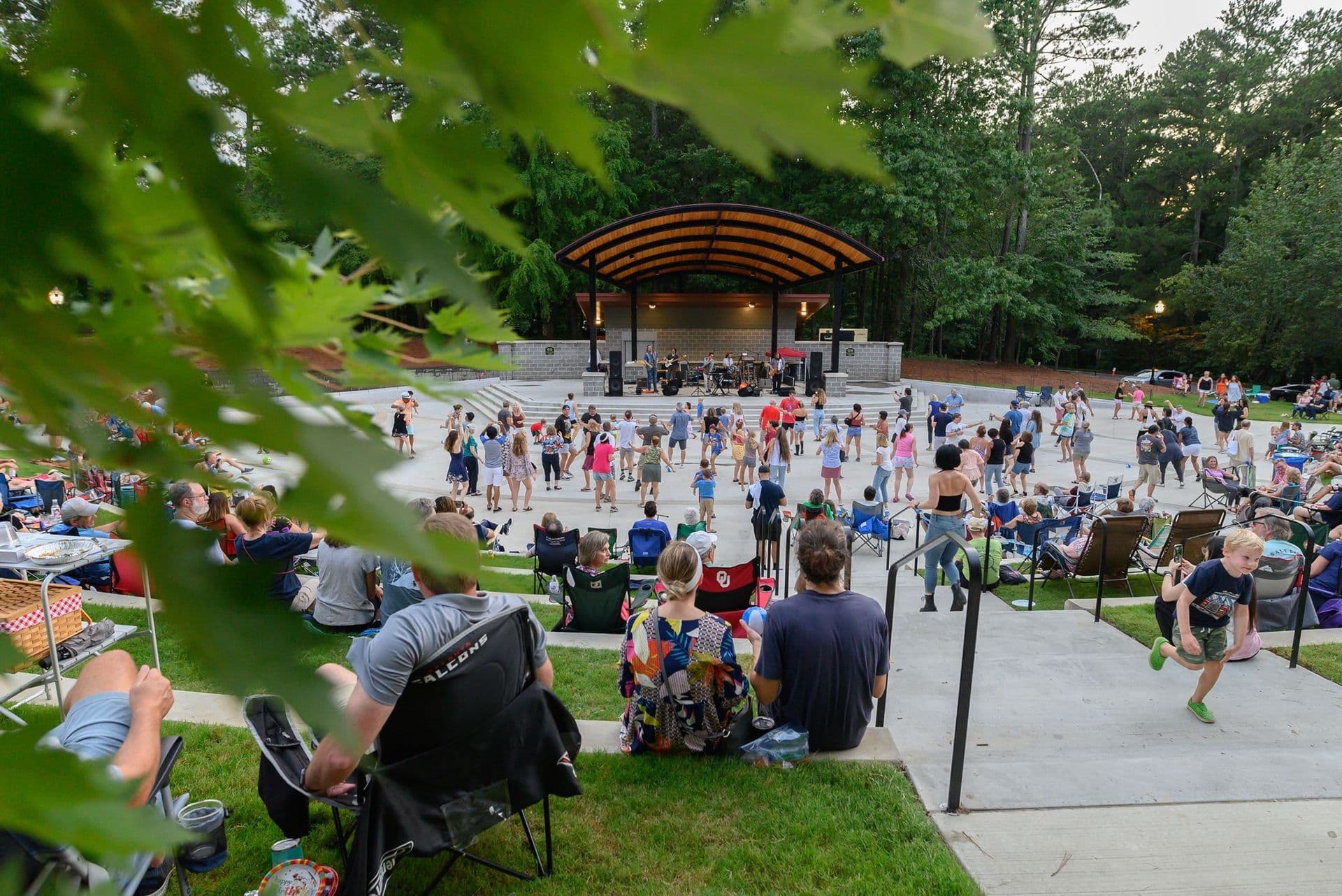 Outdoor concert scene in a park amphitheater, people sitting on lawns, children dancing near the stage. The atmosphere is lively and festive.