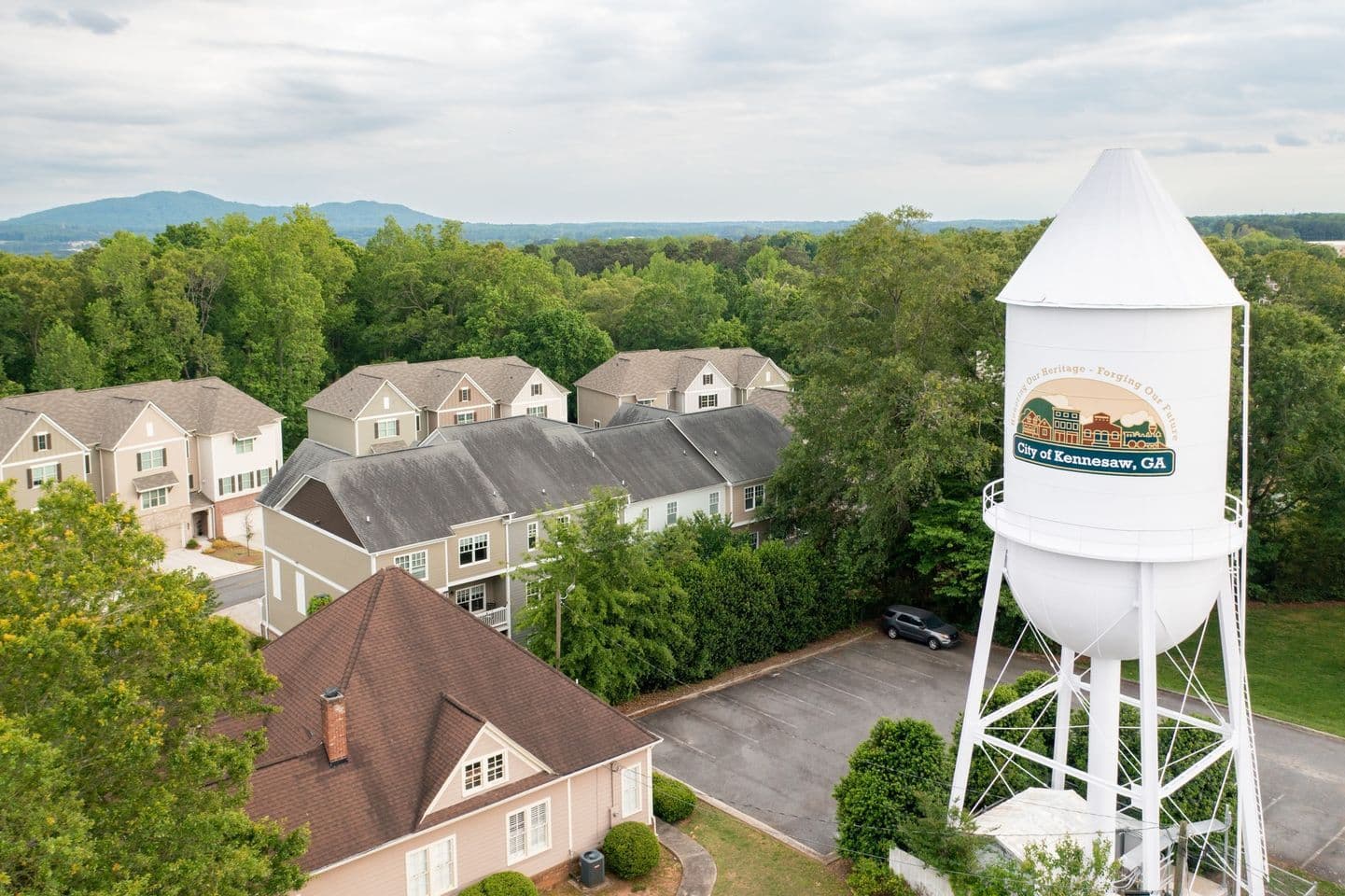 Aerial view of a residential neighborhood in Kennesaw, GA. A white water tower with the city's name stands amid lush green trees. Houses have brown and gray roofs. Mountains are visible in the distance. Quiet, suburban ambiance.