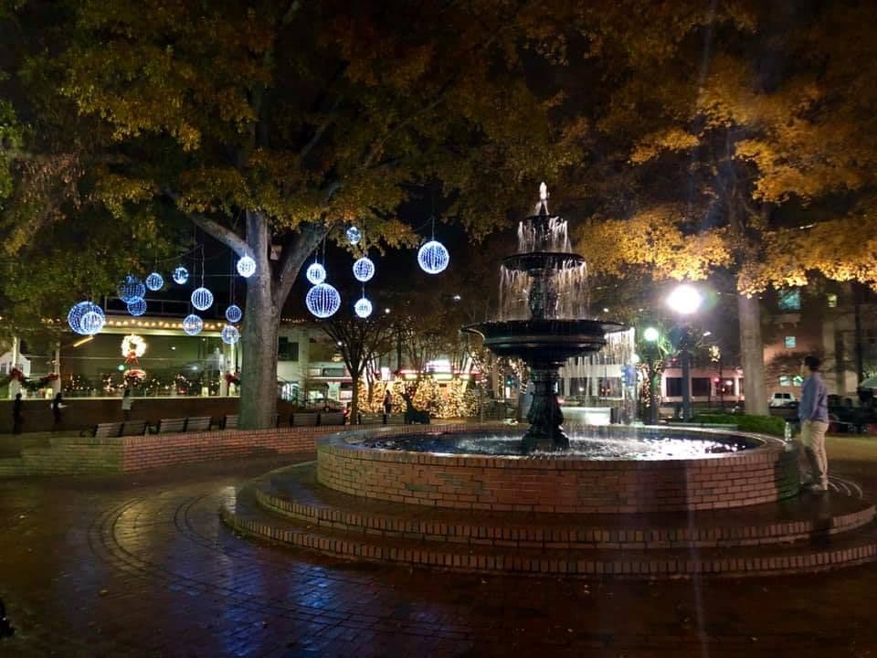A serene night scene in a park featuring a lit fountain surrounded by trees adorned with glowing spherical ornaments, creating a festive, tranquil ambiance.