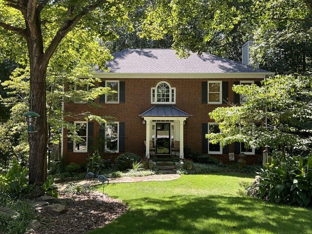 Two-story brick house with black shutters and white trim, surrounded by lush green trees and a manicured lawn, exuding a peaceful, suburban vibe.