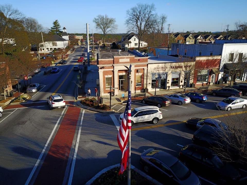 Aerial view of a small town intersection on a clear day. American flag in foreground, brick building and parked cars line streets. Trees without leaves.