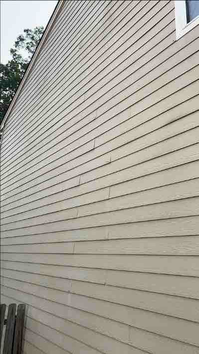 The image shows a beige, textured siding wall with visible seams and several discolored patches. In the top left, a tree is partially visible under a cloudy sky.