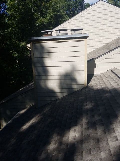 A sloped gray shingle roof with a rectangular beige chimney structure. Sunlight casts tree shadows on the roof and chimney, creating a calm tone.